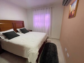 Une chambre d’hôtel avec un lit, deux oreillers, une banquette, une table de chevet, une fenêtre avec des rideaux et un tableau au mur.