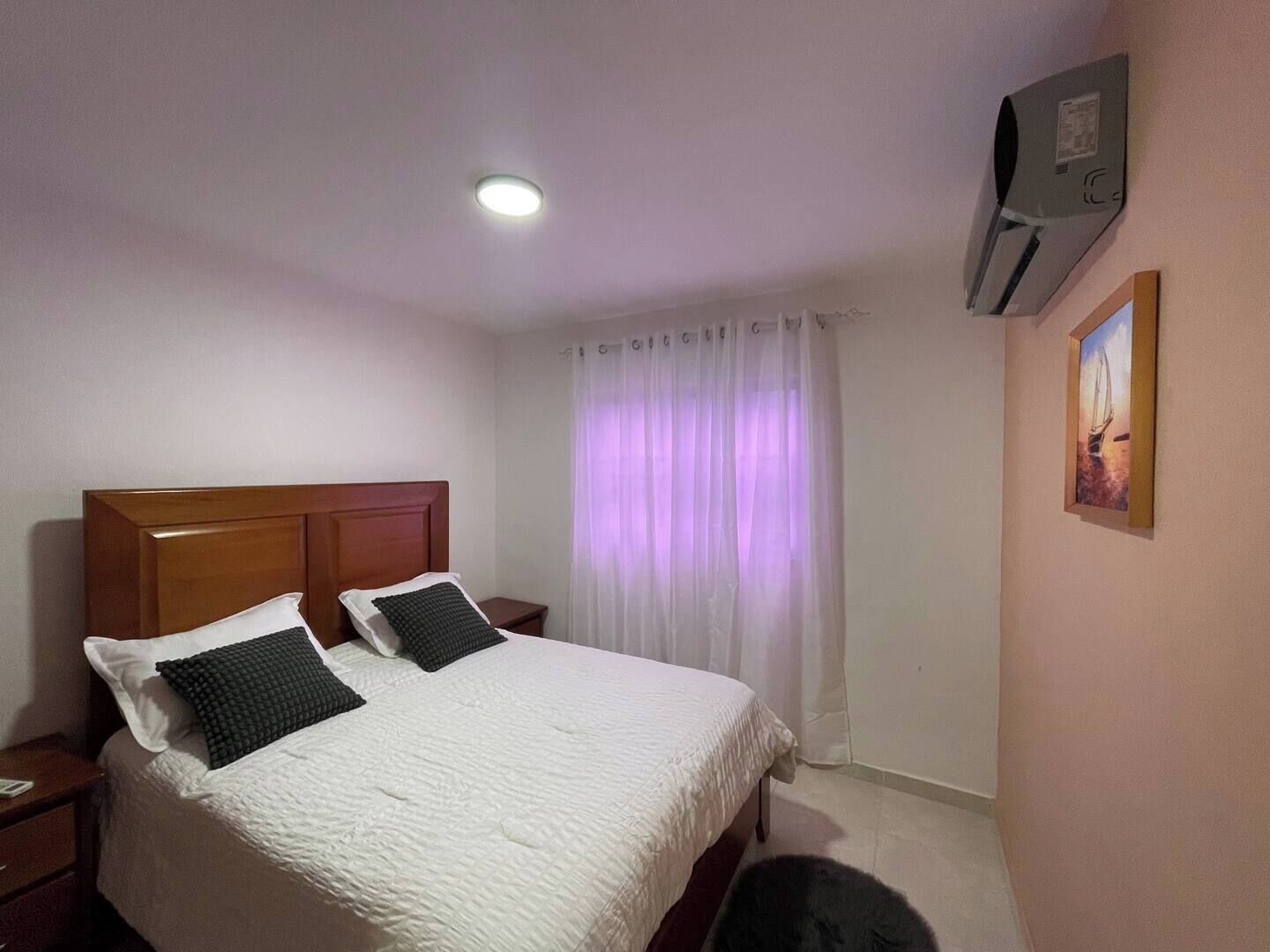 A hotel room with a wooden headboard bed, white bedding, a small bedside table, a wall-mounted air conditioner, and a window with sheer curtains.