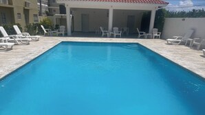 Una piscina rectangular con agua azul cristalina, rodeada de sillas de descanso blancas y una zona cubierta con asientos.