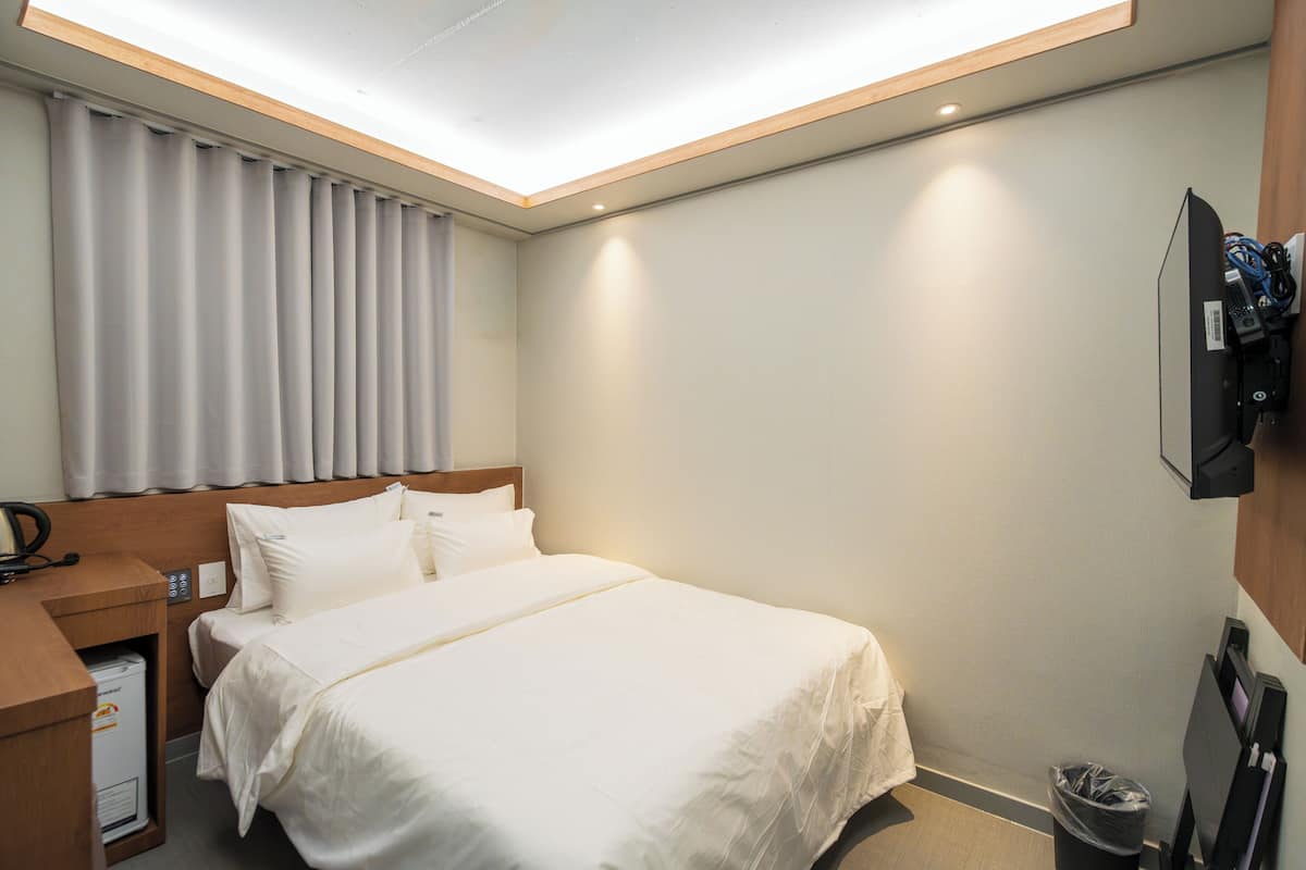 Une chambre d’hôtel comprenant un lit, un bureau avec un téléphone, un téléviseur fixé au mur et une poubelle.