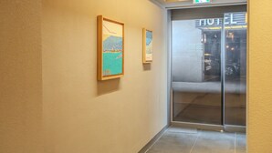 Un pasillo con dos cuadros enmarcados en la pared, una puerta de cristal que da a una habitación de hotel y una señal de salida verde encima de la puerta.