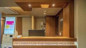Una mesa de recepción con un cartel que dice 'INFORMATION THE SQUARE HOSTEL' y un quiosco digital de información a la izquierda.