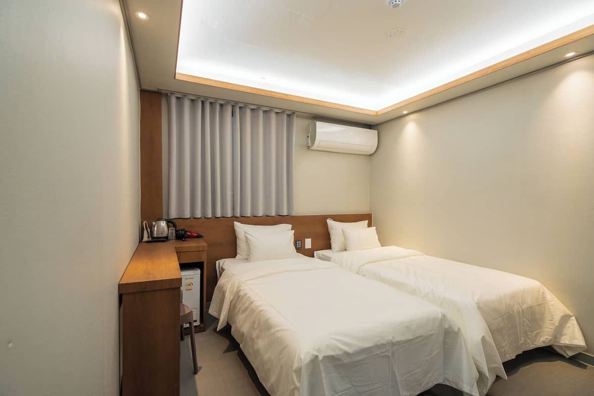 Une chambre d’hôtel avec deux lits, un bureau, une chaise, un appareil de climatisation et une fenêtre avec des rideaux.