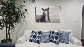 Ein Wohnzimmer mit einem beigen Sofa, drei gemusterten Kissen, einer Topfpflanze und einem gerahmten Gemälde eines Pferdes an der Wand.