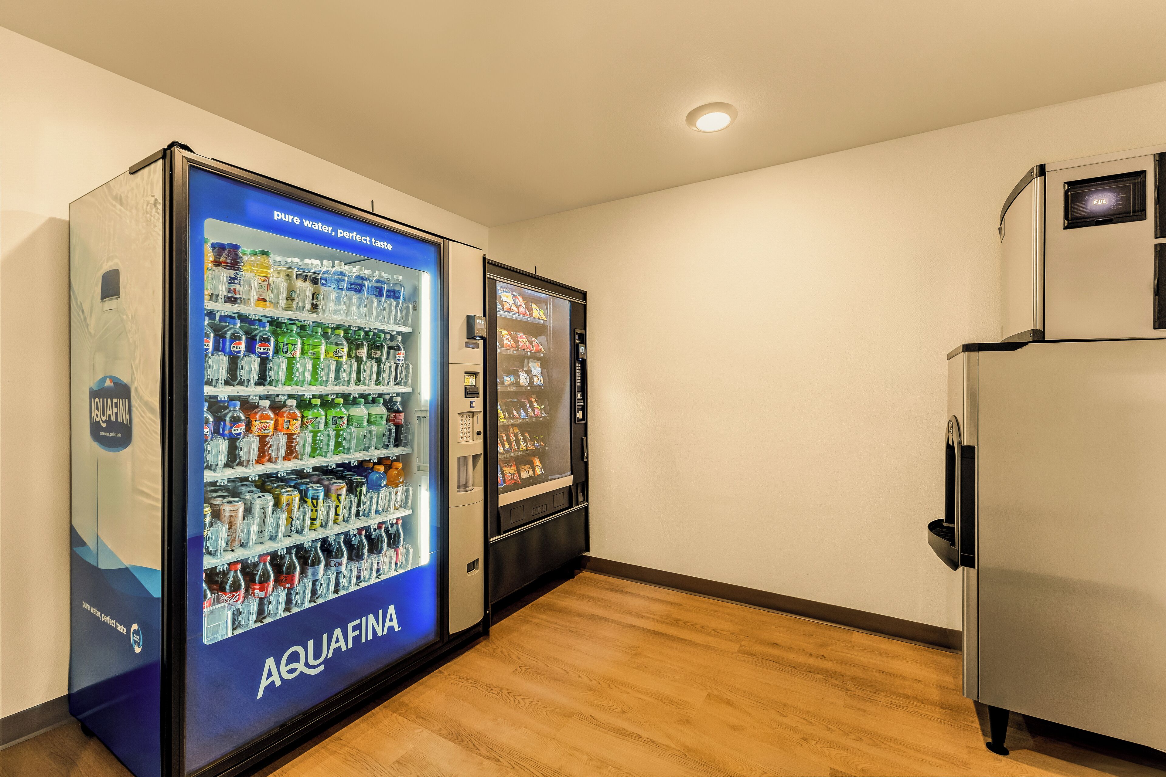 A room with a wooden floor, a refrigerator, and two vending machines labeled 'AQUAFINA'.