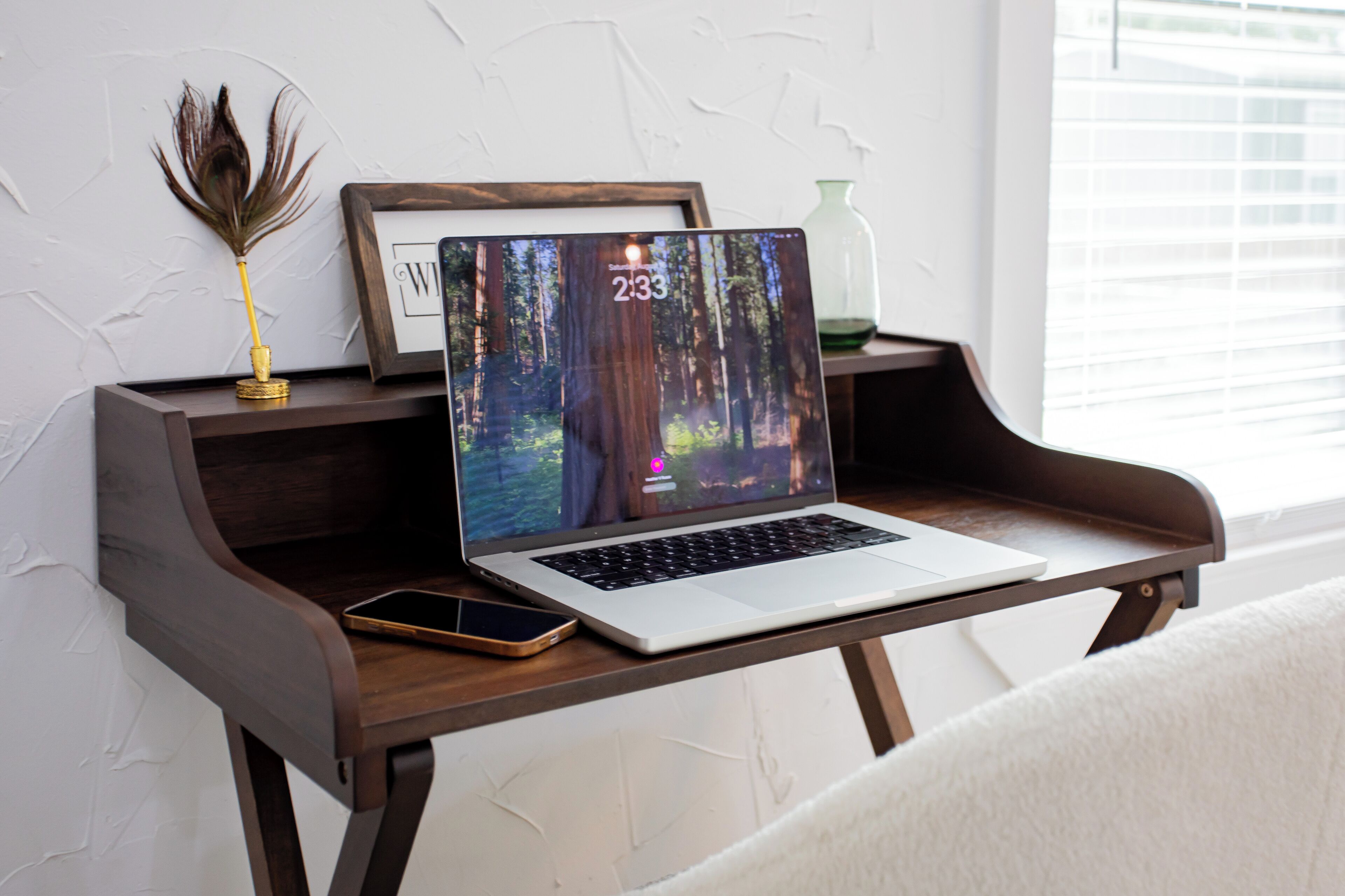 Un ordinateur portable est posé sur un bureau en bois, à côté duquel se trouvent un smartphone, un cadre photo et quelques objets décoratifs.