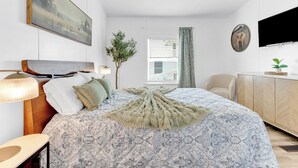 Un dormitorio con una cama grande, televisor montado en la pared, una ventana con persianas y una planta.