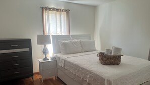 A bedroom with a bed, a dresser, a nightstand, a lamp, a window with curtains, and a basket with towels. - Charming Vacation Home Near Niagara Falls & Highmark Stadium (Depew)