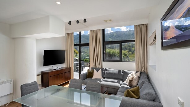 A modern living room with a flat-screen TV, a glass-top dining table, a sofa with cushions, and large windows offering a mountain view.