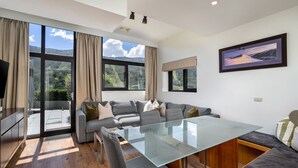 A modern living room with a glass dining table, grey sofa, and large windows offering a view of the outdoors.