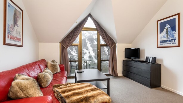 Una sala de estar moderna con un sofá de cuero rojo, una mesita, un televisor y un balcón con vistas a árboles nevados.