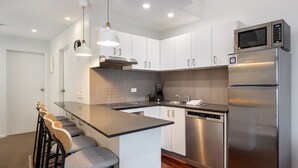 Una cocina moderna con armarios blancos, un refrigerador de acero inoxidable, un microondas y un lavavajillas integrado. Hay dos lámparas colgantes sobre la isla, y un taburete de bar con una superficie de madera.