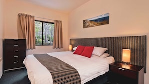 A hotel room with a bed, a dresser, a nightstand, a lamp, a painting, and a window with curtains. - Lantern 24 Balcony (Thredbo)