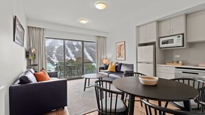 A modern hotel room with a large window offering a scenic view of mountains, a cozy seating area with a sofa and chairs, a dining table, and a kitchenette with a microwave. - Lantern 19 Deluxe (Thredbo)