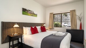 A hotel room with a large bed, two bedside tables, a nightstand, a dresser, a window with curtains, and a painting on the wall. - Lantern 19 Deluxe (Thredbo)