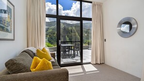 Ein Zimmer mit einer Couch, gelben Kissen, einem runden Spiegel und einer Schiebeglastür, die auf eine Terrasse mit Tisch und Stühlen führt.