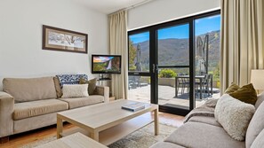 A modern living room with a beige sofa, a wooden coffee table, and a flat-screen TV. The room has a large sliding glass door leading to an outdoor patio with a table and chairs.