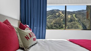 A bed with red and beige pillows, a view of a mountainous landscape through a window with blue curtains. - Lantern 12 Balcony - views from every window (Thredbo)