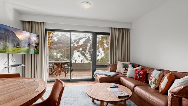 A modern living room with a large TV, a brown leather sofa, a wooden coffee table, and a dining area with a wooden table and chairs.