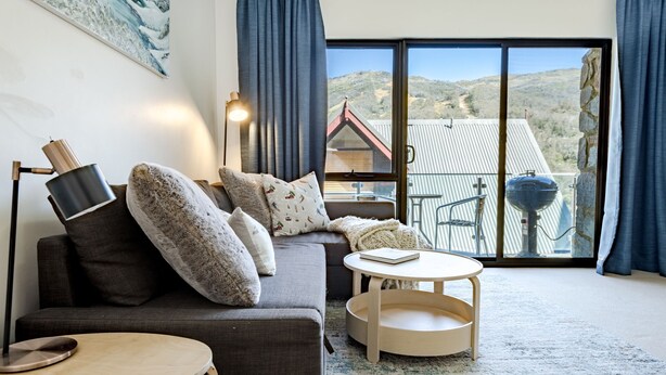 A cozy living room with a sofa, a small round table, and a view of a mountainous landscape through a large sliding glass door. - Lantern 08 Balcony (Thredbo)