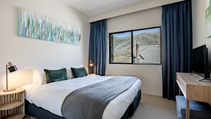 A hotel room with a large bed, a desk with a TV, a window with a view of a mountainous landscape, and a painting on the wall. - Lantern 08 Balcony (Thredbo)