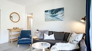 A modern living room with a blue armchair, grey sofa, and a white coffee table. A painting of a snowy landscape hangs above the sofa. - Lantern 08 Balcony (Thredbo)