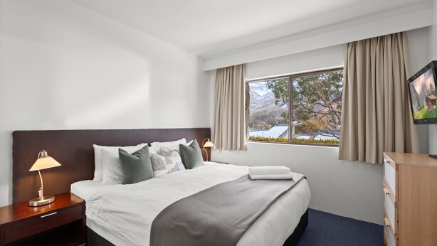 Ein Hotelzimmer mit Bett, Nachttisch, Lampe, Fernseher und einem Fenster mit Vorhängen.