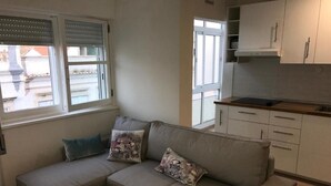 A living room with a grey sofa, two pillows, a small white table, and a kitchen area with white cabinets and a black countertop.