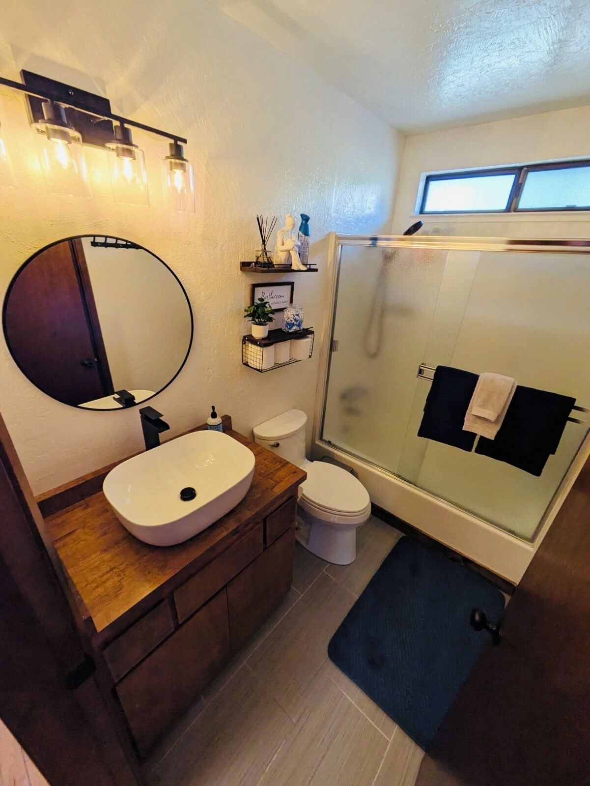 A modern bathroom with a round mirror, wooden vanity, white sink, toilet, and glass-enclosed shower.