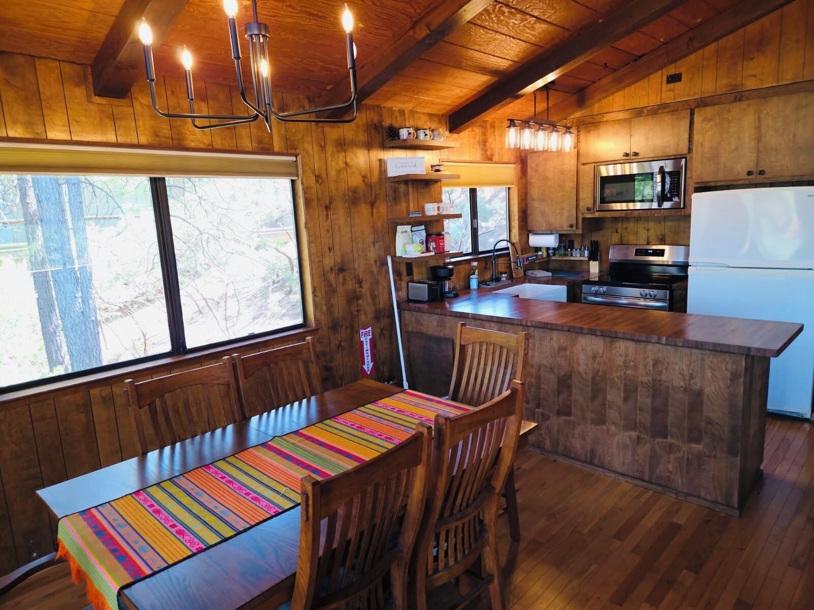 Une cuisine rustique aménagée dans une cabane en bois, avec une table à manger, des chaises, une grande fenêtre et un îlot central doté d’un évier et d’une cuisinière.
