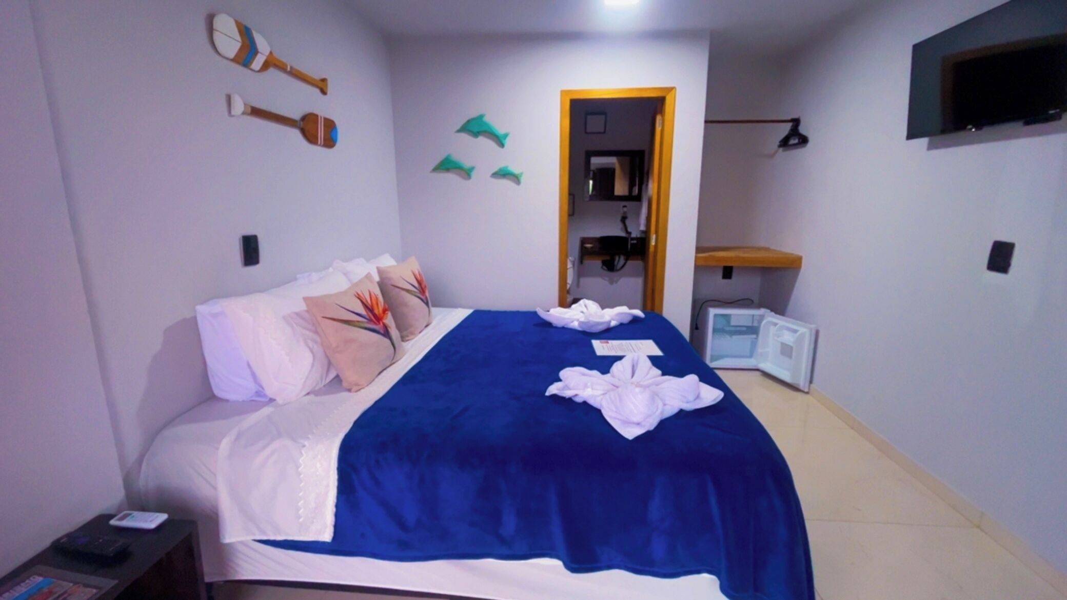 Una cama bien hecha con una manta azul y almohadas decorativas, un televisor montado en la pared y un escritorio pequeño con una lámpara.