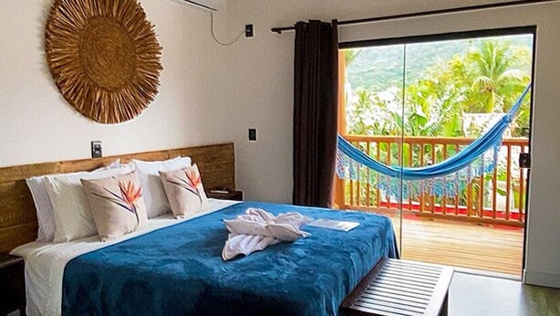 A bedroom with a bed, a bench, a wall-mounted air conditioner, a woven wall decoration, and a view of a balcony with a hammock. - Pousada Amaris (Penha)