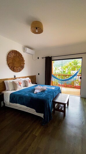 Un dormitorio con cama, un banco, aire acondicionado de pared, un adorno de pared tejido y vistas a un balcón con hamaca.