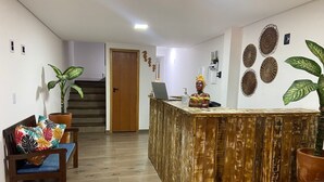 A reception area with a wooden reception desk, a laptop, and a decorative statue. There are two chairs with colorful cushions and potted plants. - Pousada Amaris (Penha)