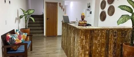 Zona de recepción con un mostrador de madera, una computadora portátil y una estatua decorativa. Hay dos sillas con cojines de colores y plantas en macetas.