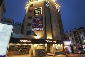 Un bâtiment avec une enseigne indiquant « TVOSOME STAY » et une banderole annonçant « Grande ouverture en janvier 2024 ». Il y a un espace de stationnement avec une enseigne « PARKING » et une enseigne « TVOSOME STAY » à l’entrée.