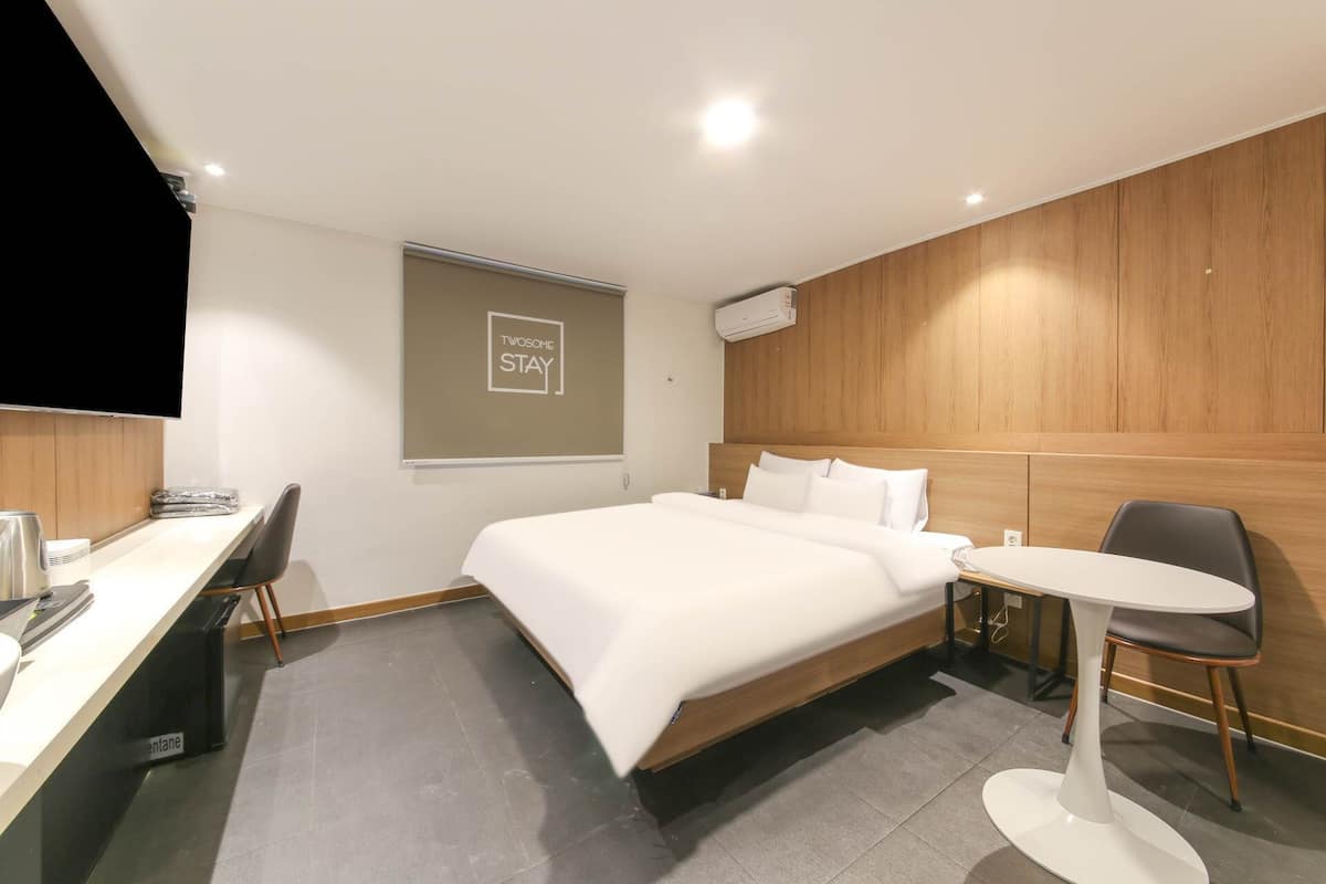 A modern hotel room with a bed, a desk, a chair, a small table, and a wall-mounted air conditioner.