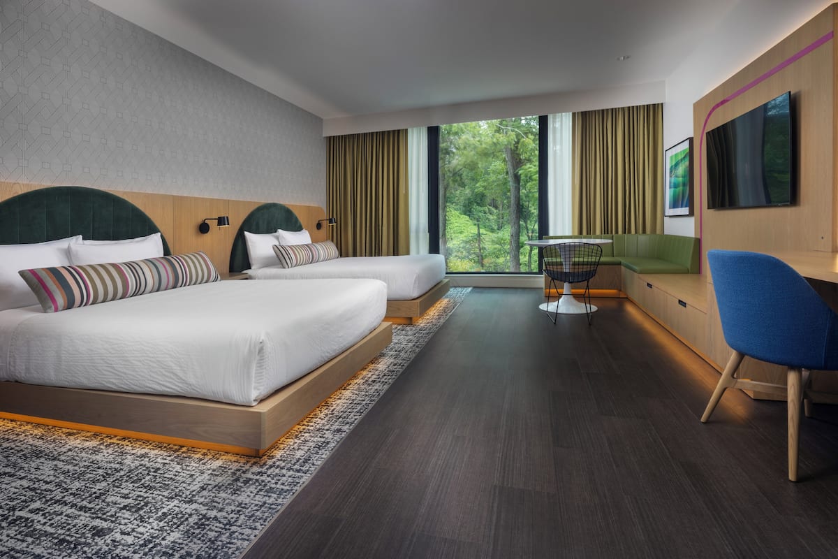 Ein modernes Hotelzimmer mit zwei Betten, einem Schreibtisch, einem Stuhl und einem großen Fenster mit Blick auf Grünflächen.
