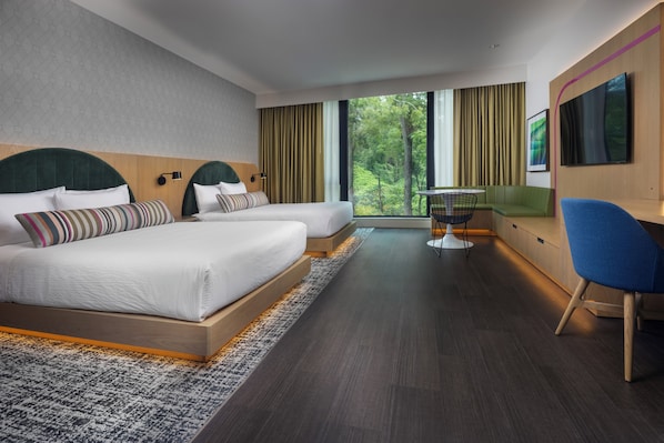 A modern hotel room with two beds, a desk, a chair, and a large window offering a view of greenery.