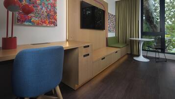 Une chambre moderne avec un bureau en bois, une chaise bleue, un téléviseur fixé au mur, une petite table avec une chaise et une grande fenêtre avec des rideaux.