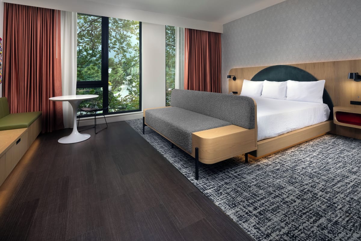 Ein modernes Hotelzimmer mit Schlafsofa, einem kleinen Tisch, einem Sessel und einem großen Fenster mit Vorhängen.