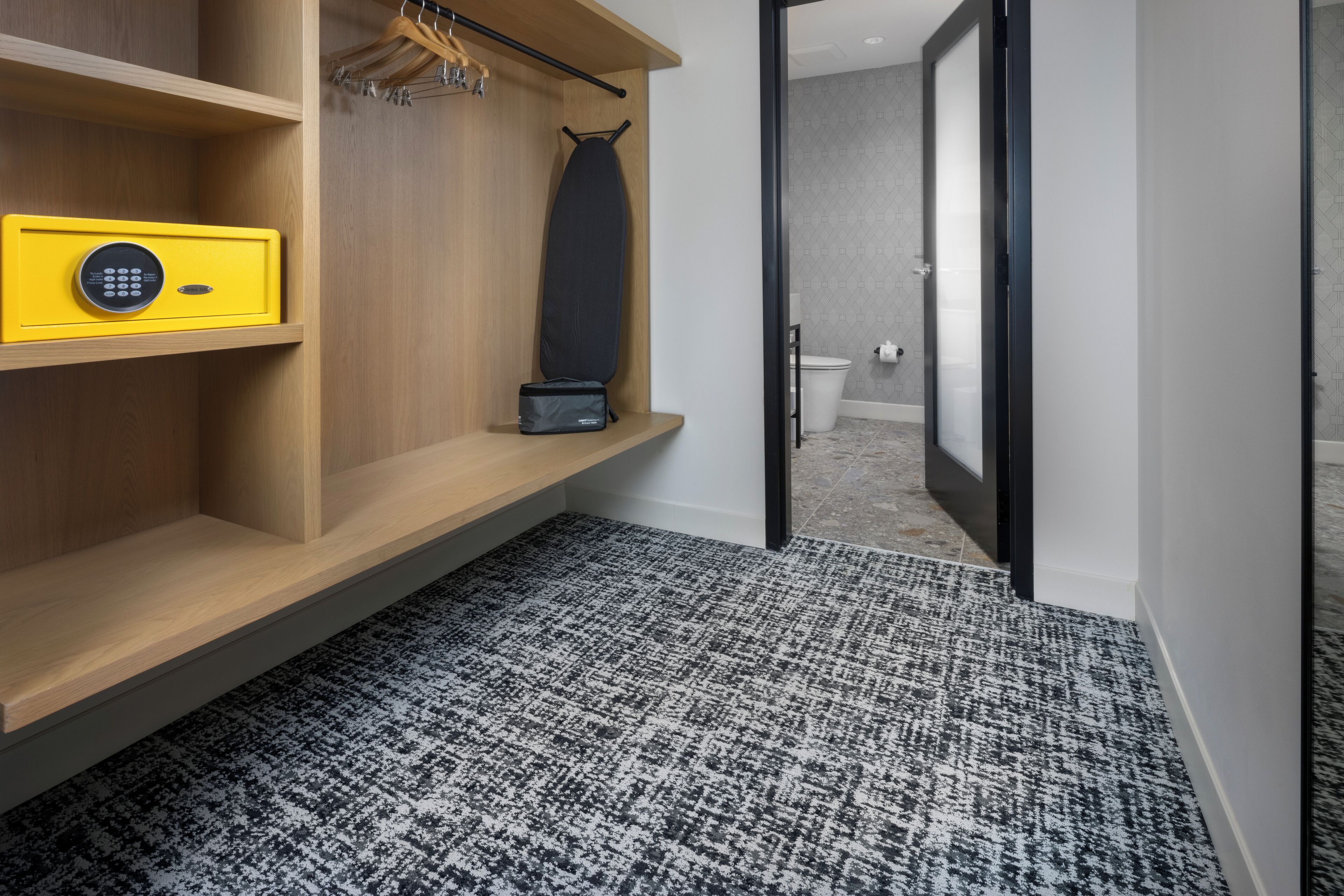 A hotel room with a wooden shelf, a yellow safe, a black bag, and a patterned carpet.