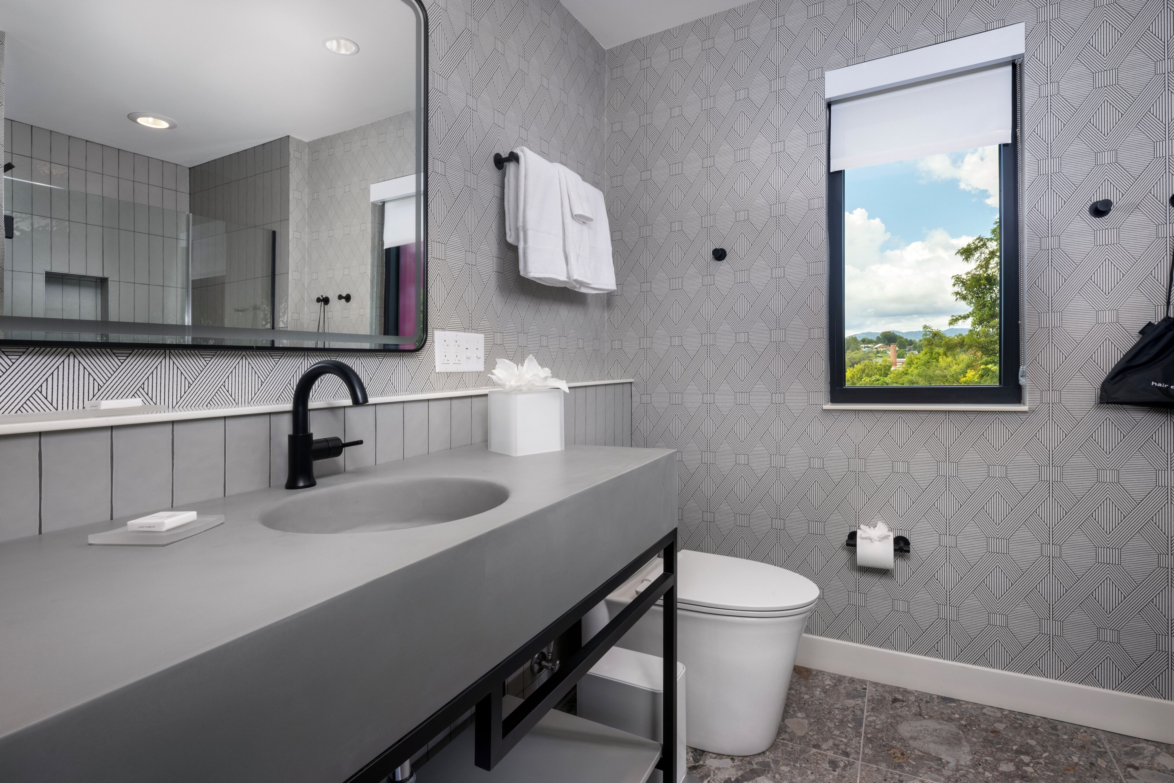 Un baño moderno con pared de azulejos grises, un espejo grande, un lavamanos negro, un inodoro blanco y una ventana con vista a follaje.