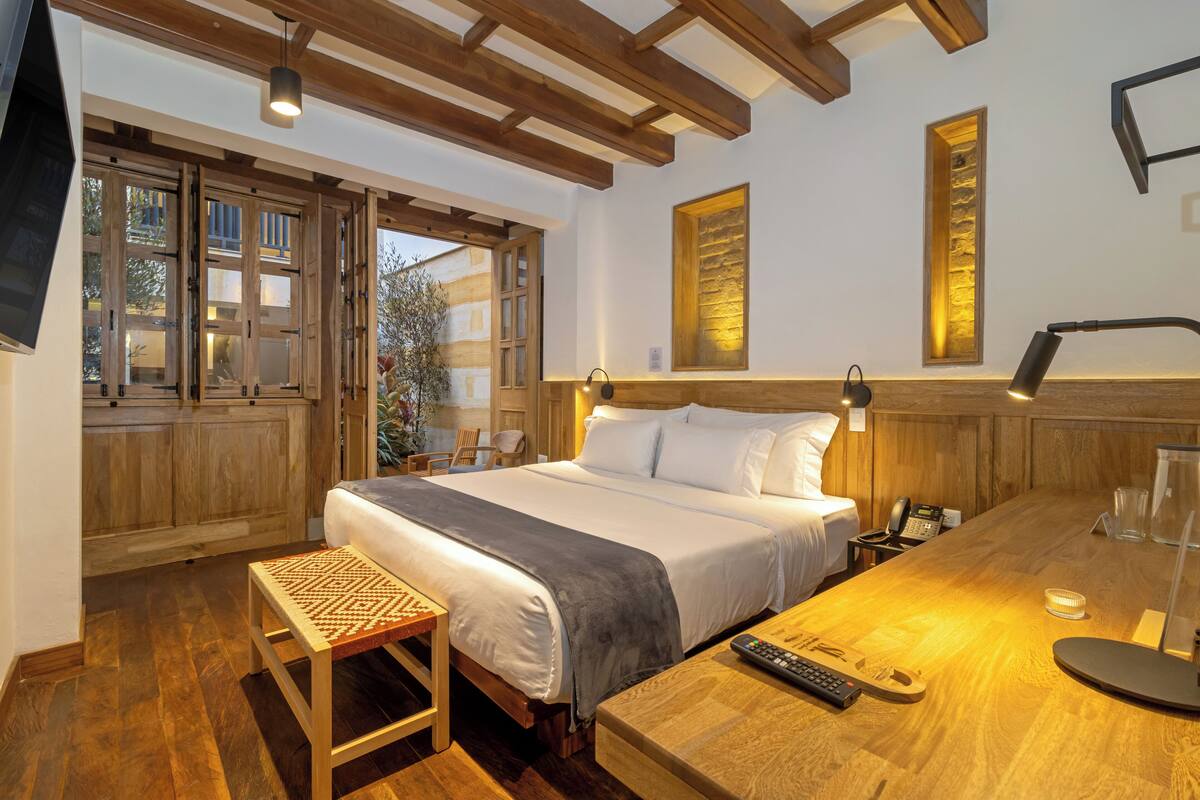 A bedroom with a large bed, wooden ceiling beams, a desk with a lamp, and a window with a view of the outside.