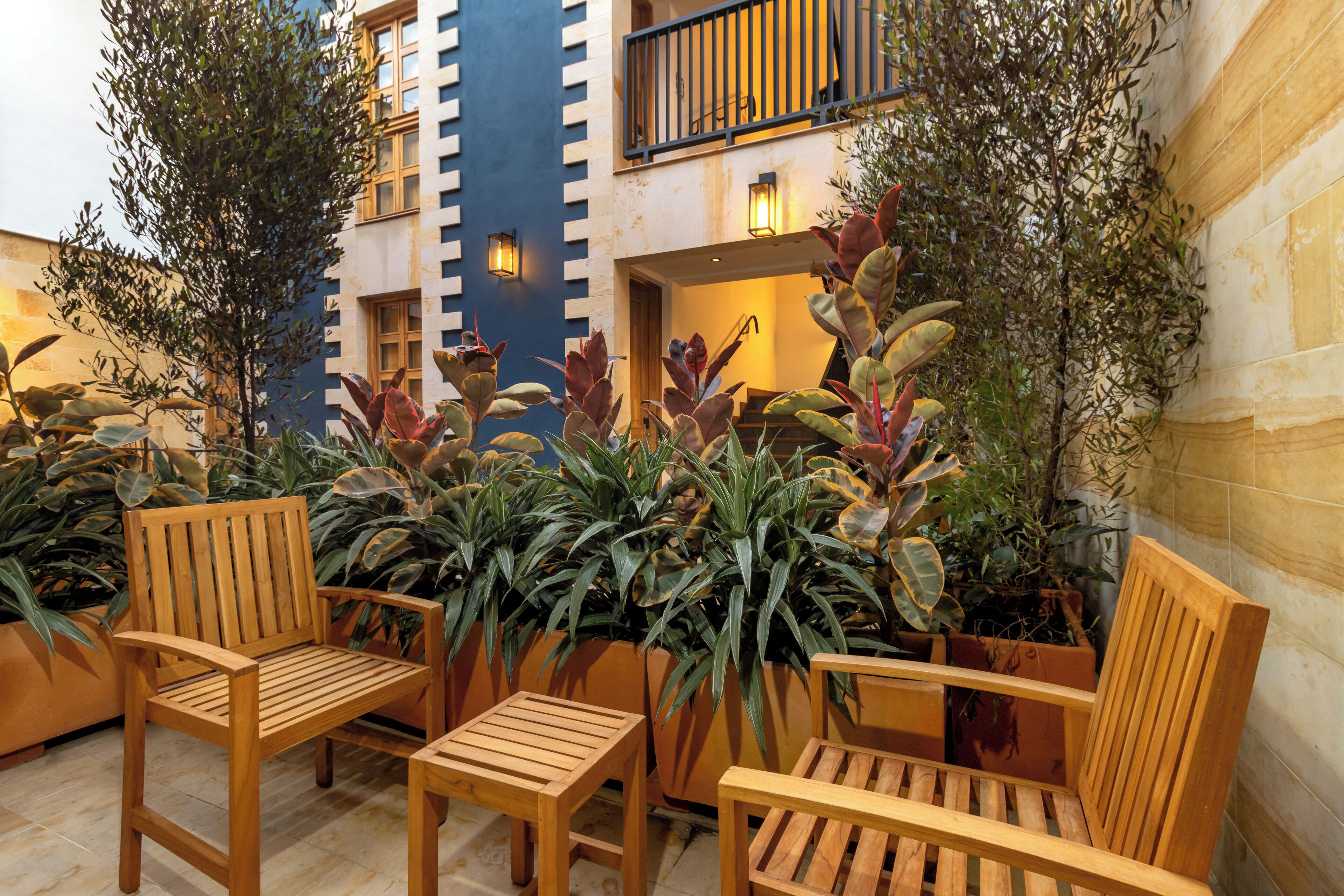 Un patio con sillas de madera y plantas en macetas, un edificio con fachada azul y un balcón con barandillas.