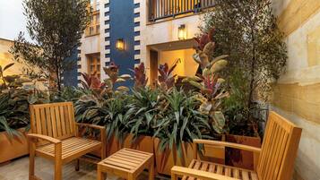 A courtyard with wooden chairs and potted plants, a building with a blue facade, and a balcony with railings.