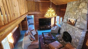 Ein gemütliches Blockhaus-Interieur mit Kamin aus Stein, Ledersofas, einem Fernseher und einer Holzbalkendecke.