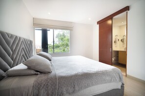 Quarto moderno com uma cama grande, cabeceira com padrão geométrico, janela com vista para vegetação e porta deslizante que dá para um banheiro.