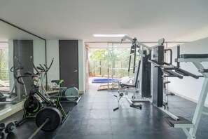 Ginásio com equipamentos de exercício, espelho e vista para um jardim.
