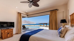 Habitación de hotel con cama, televisor, ventilador de techo y vista a la playa y al océano.
